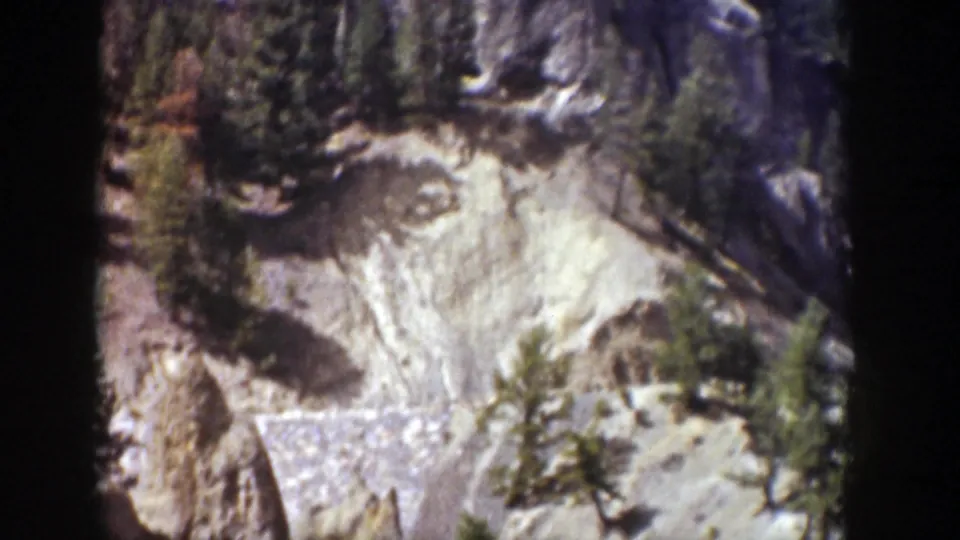 Wyoming home-movie frame with pale cliff textures, scattered evergreens, and strong sunlight on the rocky slope.