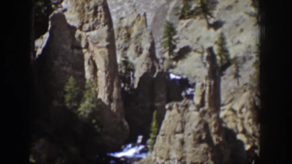 Final Wyoming 8mm frame with tall rock spires, scattered evergreen trees, and a shadowed opening or recess in the cliff.