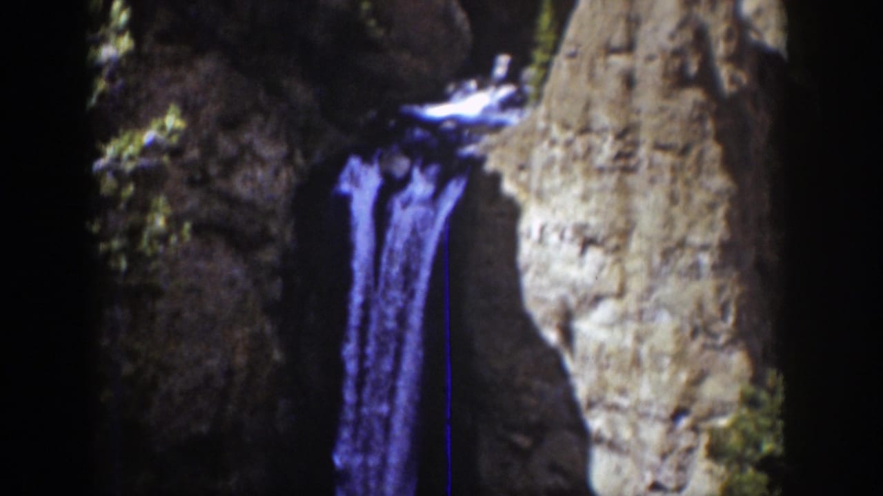 1939 Wyoming scenic frame with blue-purple waterfall water dropping beside pale canyon rock and shadowed cliff face.
