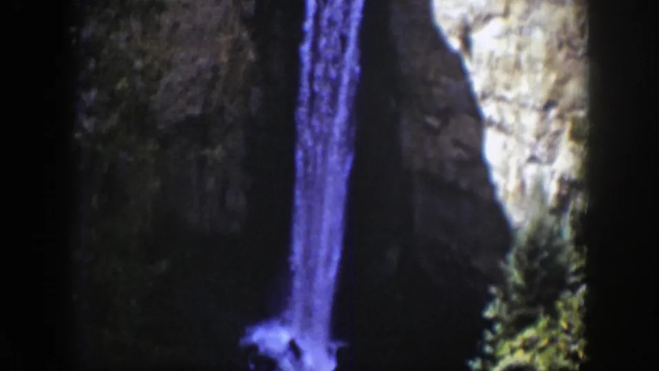 1939 Wyoming home-movie frame showing a tall mountain waterfall, foamy splash at the base, and evergreens along the canyon wall.