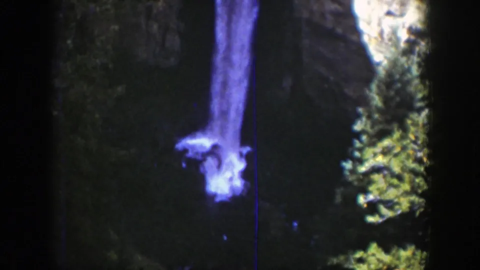 1939 Wyoming 8mm frame of a waterfall against a shadowed cliff, bordered by evergreens and pale rock at the canyon edge.
