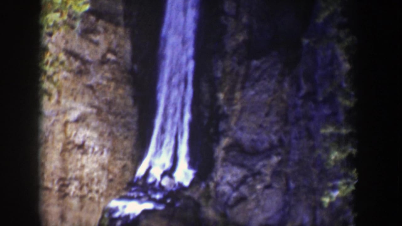 Narrow waterfall dropping between dark rock walls with green bushes at the upper edge.