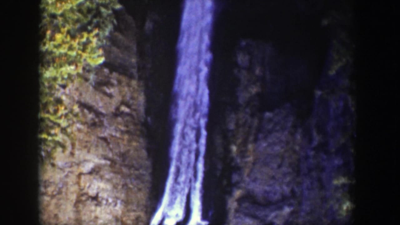 Vintage 8mm Wyoming frame of a narrow waterfall flowing down a rocky chute, with dark cliff shadows and foliage near the top.