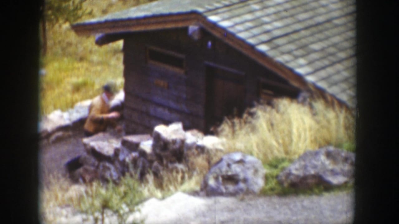 1939 Wyoming home-movie frame of a man near a small wooden cabin, with a stone wall, tall grass, and large rocks in view.