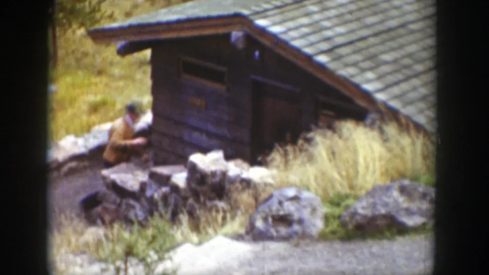 1939 Wyoming home-movie frame of a man near a small wooden cabin, with a stone wall, tall grass, and large rocks in view.