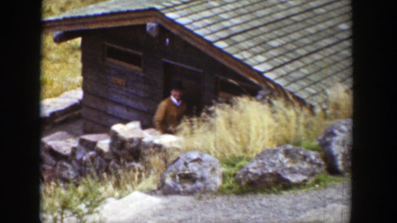 Man standing or emerging near the door of a small wooden cabin.