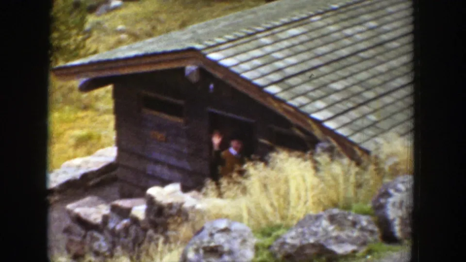 Two dark human figures appear near the doorway of a wooden cabin, partly obscured by tall grass and the film frame edge.