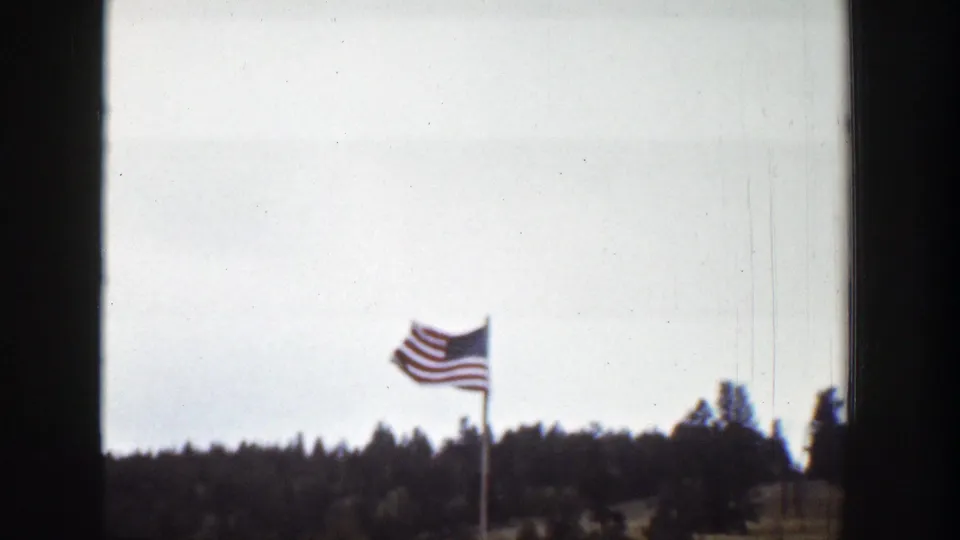 American flag waves against a cloudy sky over a forested Wyoming landscape, seen through dark frame or window edges in 1939 8mm footage.