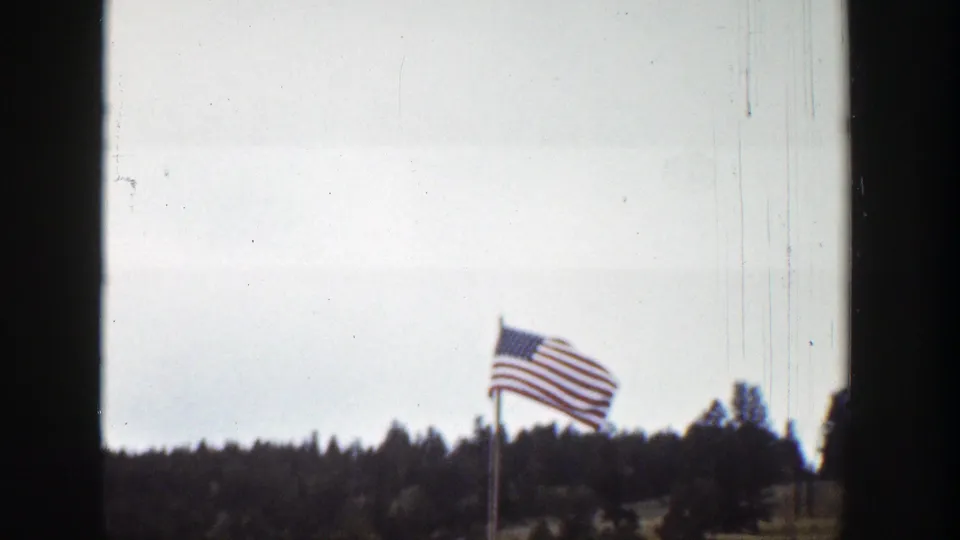 American flag on a tall pole waves over a pine-covered landscape in Wyoming, with the scene partially bordered by a dark vertical frame edge.