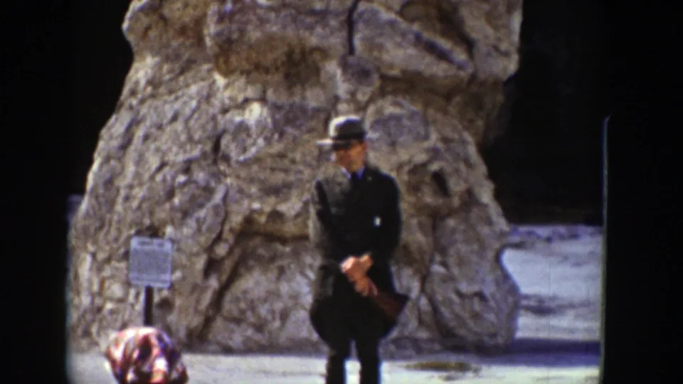 1939 Wyoming frame showing a man in a dark suit and hat standing with hands clasped before a large rock formation.
