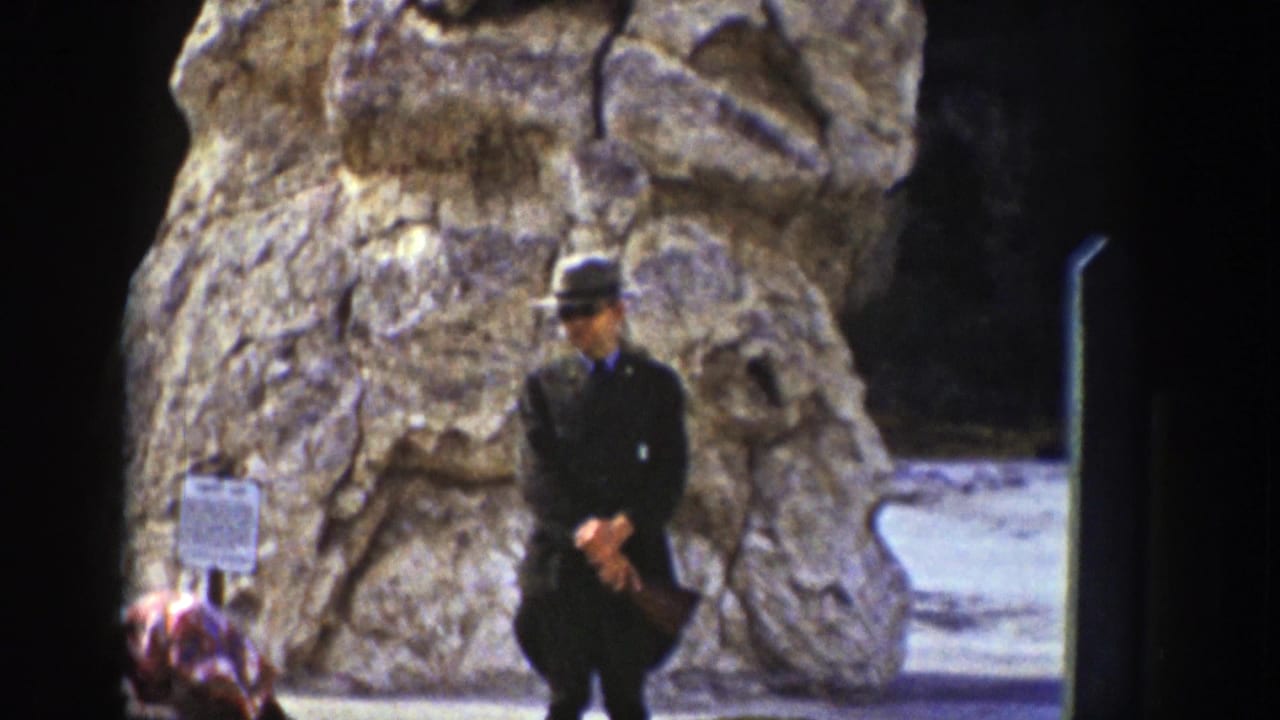 Man in dark suit and hat standing before a large carved stone monument with a small sign nearby.