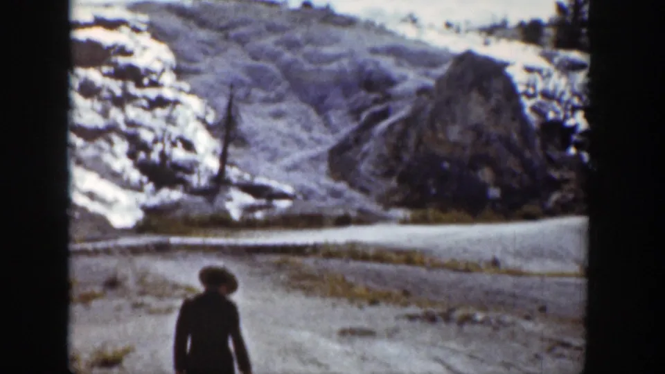 1939 Wyoming frame with a lone walker on a dirt path below snow-streaked rocky hills and sparse trees.