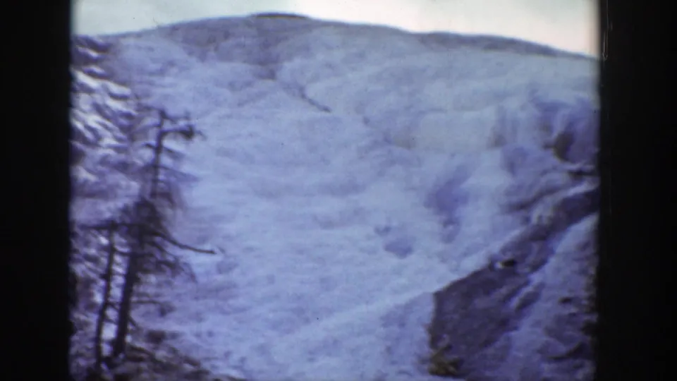 A broad pale snow-covered hillside rises behind sparse dark trees and exposed rock in this Wyoming 1939 8mm winter view.