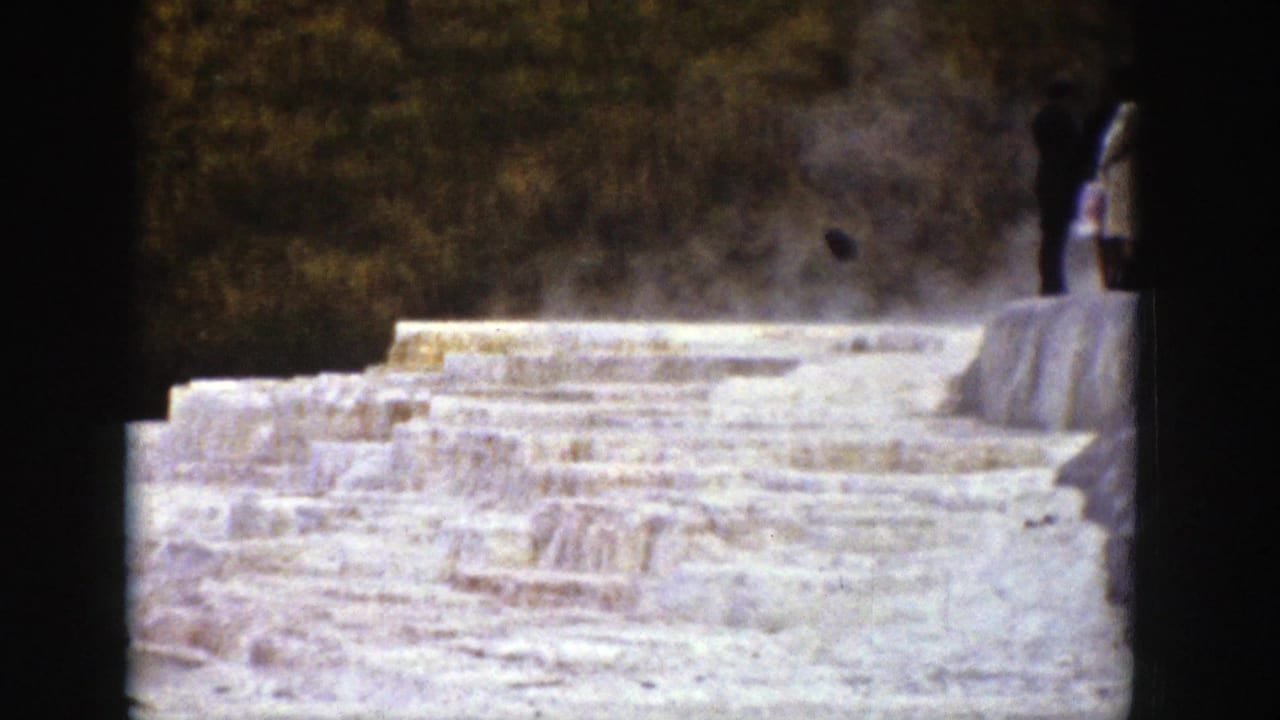Aged 8mm view of steaming mineral terraces in Wyoming, with a partial visitor visible at the right side of the frame.