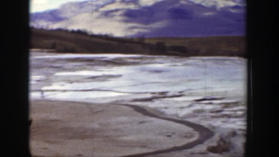 1939 Wyoming home-movie frame with a curved foreground track, pale dry terrain, dark foothills, and distant mountain ridges.