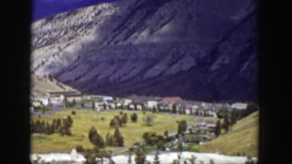 Wyoming mountain-town view with clustered buildings, open grass, trees, and steep rocky slopes behind them.