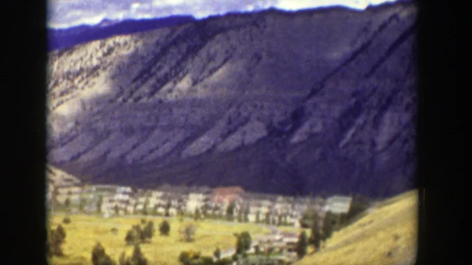Archival 8mm frame showing a closer view of a valley settlement with trees, light buildings, and a rugged mountain backdrop.
