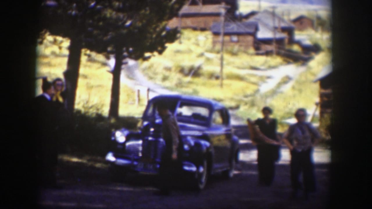 Adults stand near a dark vintage sedan on a shaded dirt road with cabins behind.