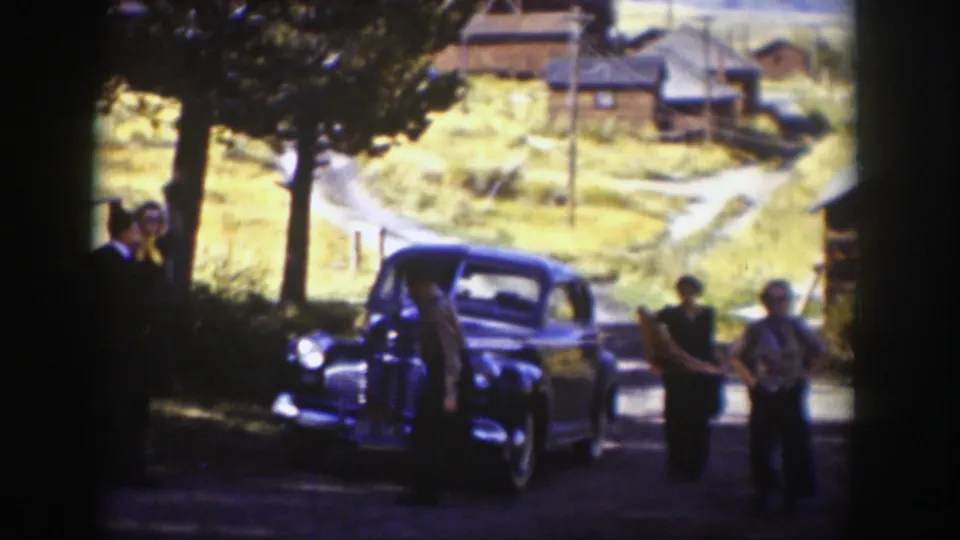 In Wyoming, 1939, adults gather beside a dark 1930s sedan on a shaded road, with wooden buildings and dry hillside in the background.