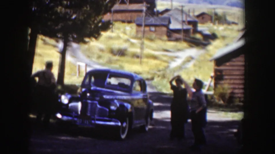 Several adults stand beside a dark 1930s sedan on a Wyoming road, with cabins, trees, and a sunlit hillside behind them.
