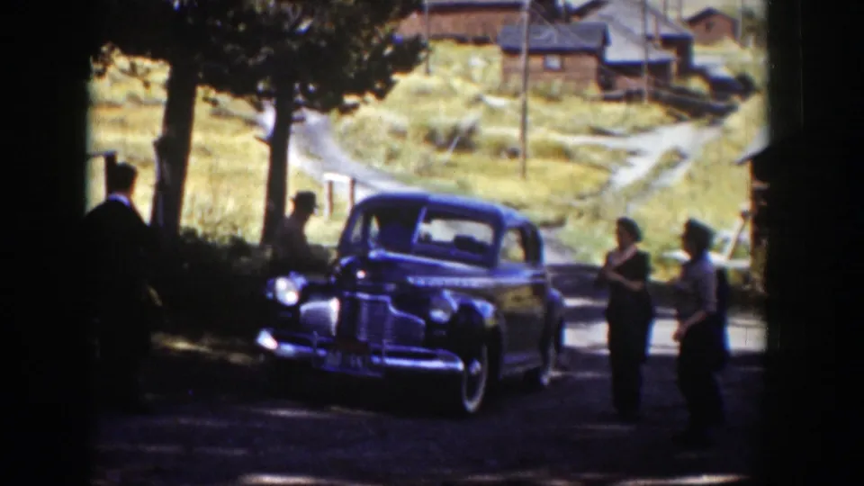 Adults in period clothing stand and converse near a parked sedan in Wyoming, 1939, with cabins, trees, and dry hills visible beyond.