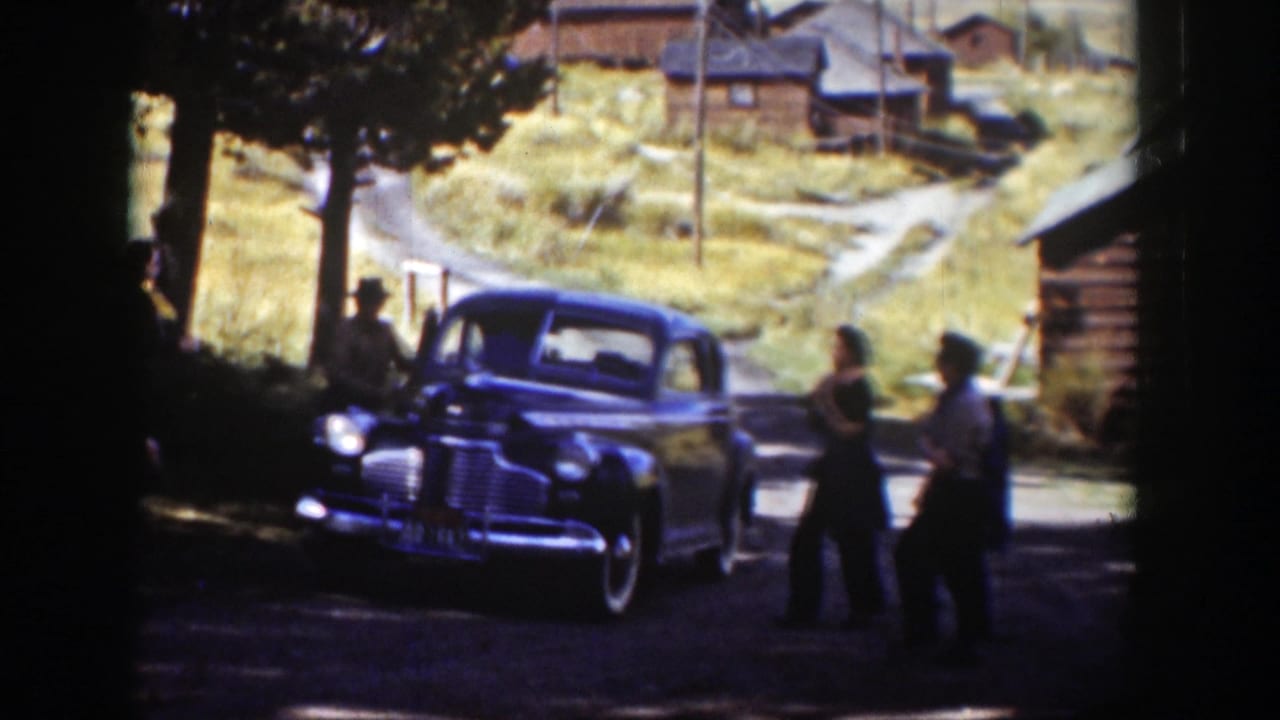 In rural Wyoming home-movie footage from 1939, adults gather beside a dark sedan near cabins, trees, dirt lanes, and a grassy hillside.