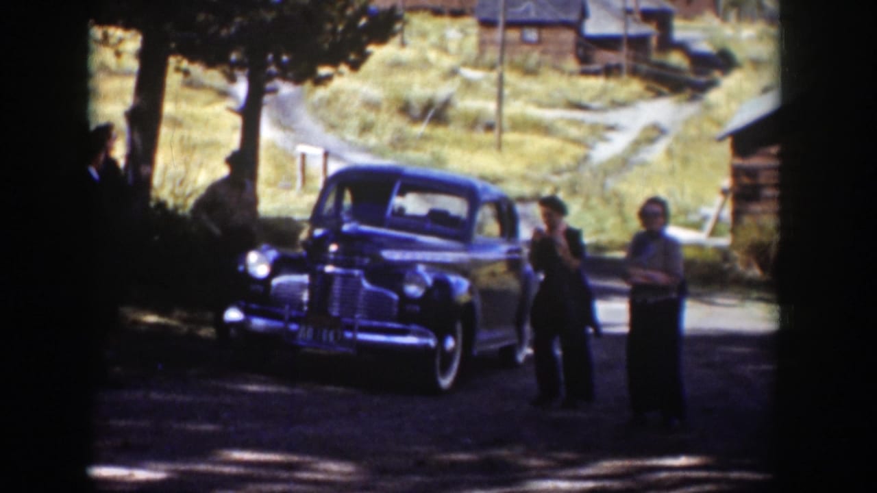 A parked sedan sits on a dirt road while several adults stand and talk near trees and cabins.