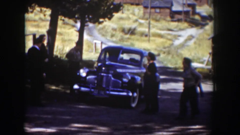Adults walk and stand near a dark 1930s-style sedan in a Wyoming settlement, with shaded foreground, cabins, dirt roads, and dry grass visible.