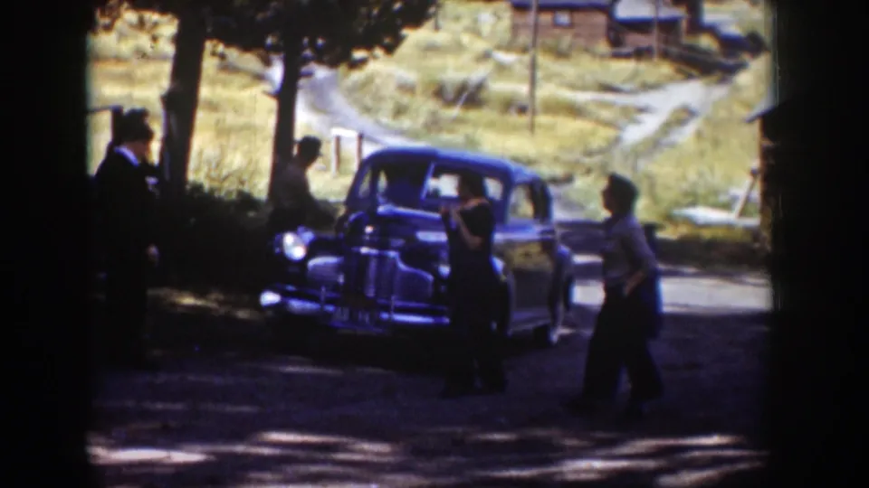 Two adults stand by the front of a parked sedan as another person walks across the shaded road in a rural Wyoming settlement scene.