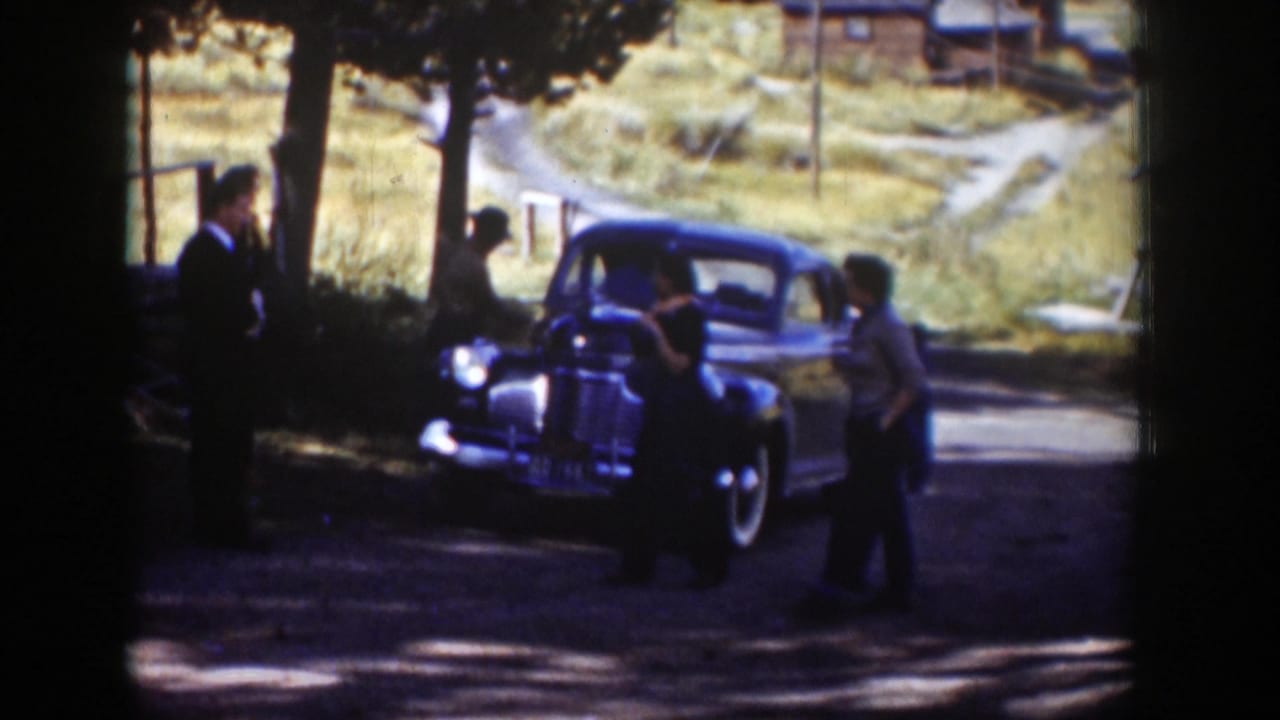 Adults gather beside a dark vintage car on a shaded rural road.