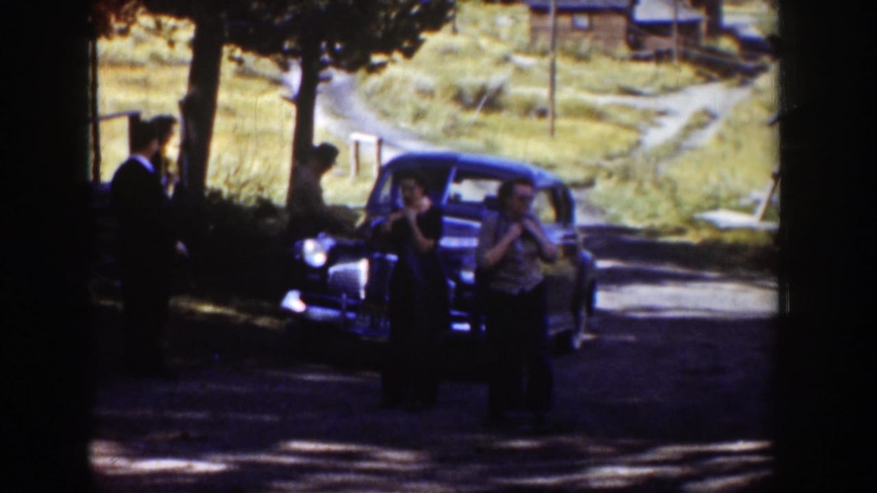 Adults in period dress gather around a dark car in rural Wyoming, with trees, grass, and a dirt road visible behind them.