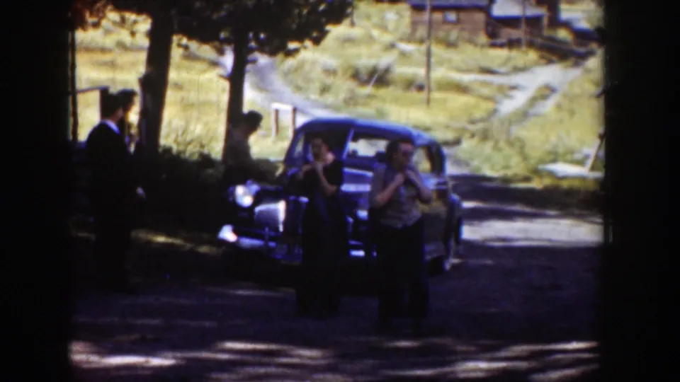 Adults in period dress gather around a dark car in rural Wyoming, with trees, grass, and a dirt road visible behind them.