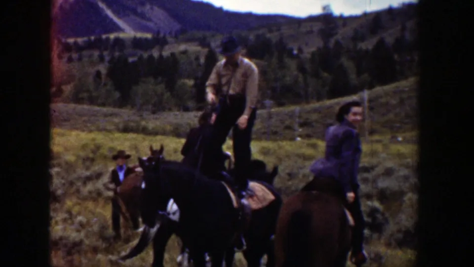 Riders on horseback move through open Wyoming hills, with grassy slopes, scattered trees, and distant mountains visible.