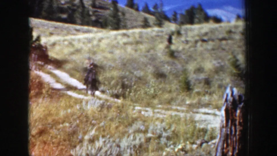 Two riders appear along the hillside trail in 1939 Wyoming home-movie footage, with a cut tree stump near the lower right foreground.