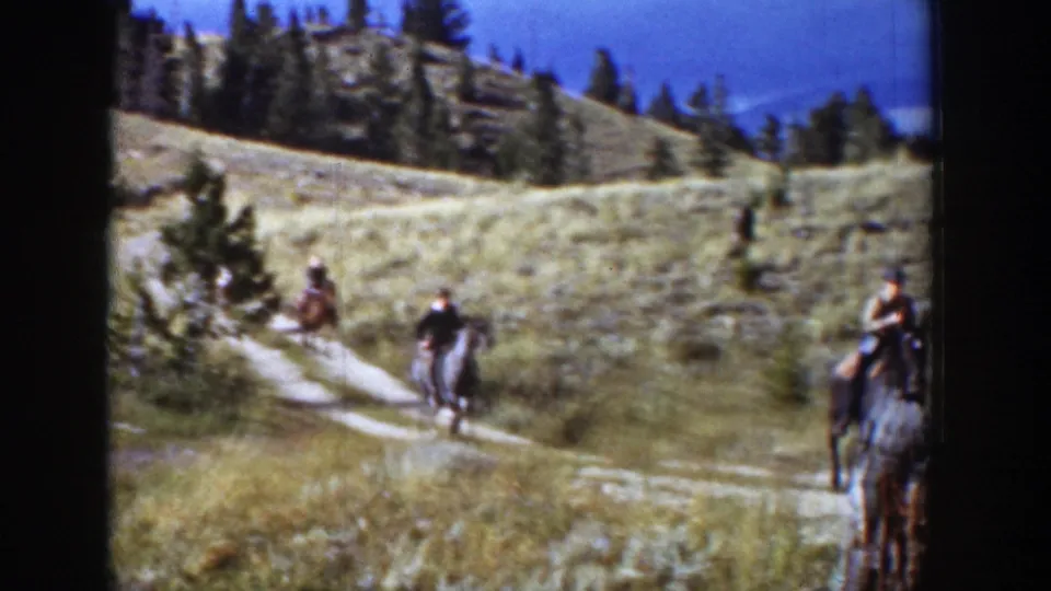 Several horseback riders pass through the sunlit Wyoming hillside, with a winding path, dry grass, trees, and a foreground stump visible.