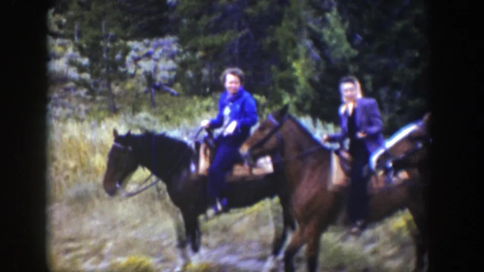 Two riders, including one in a blue jacket, are seen on horseback near a forest edge in the Wyoming riding footage.