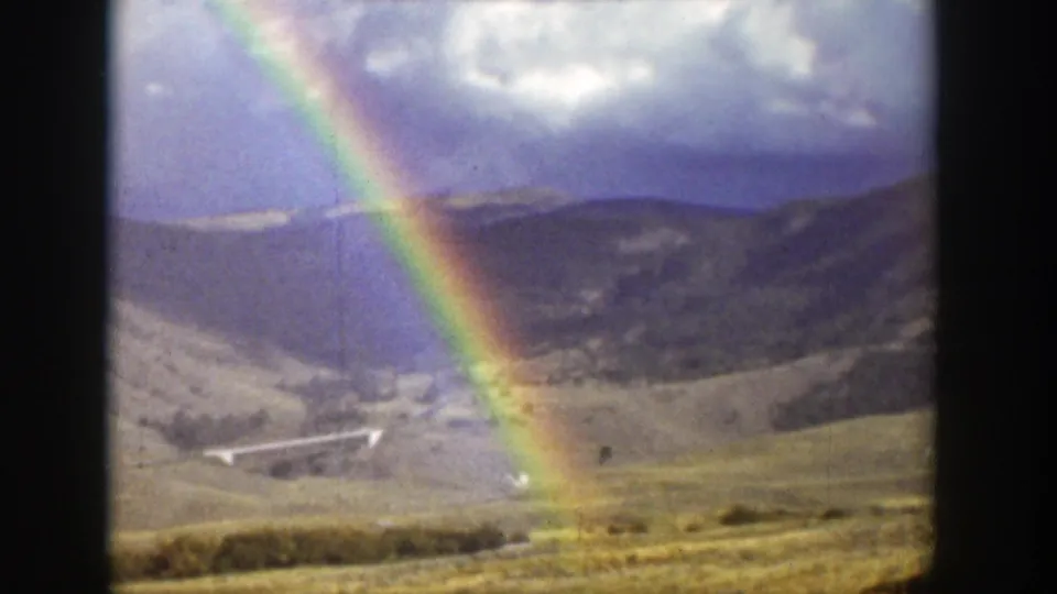 The rainbow reaches toward a grassy Wyoming hillside, with distant mountains, clouds, and 8mm grain visible.