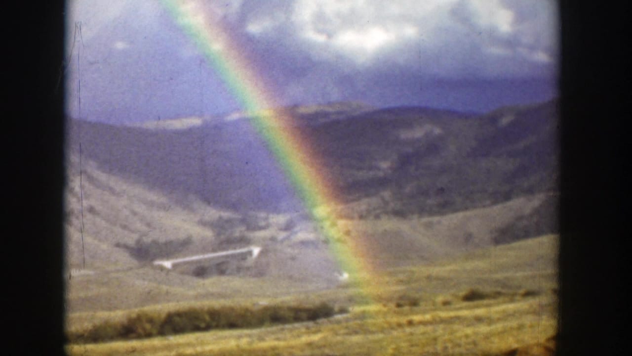 Rainbow over dry hills and a cloudy sky with a small distant bridge or road feature.