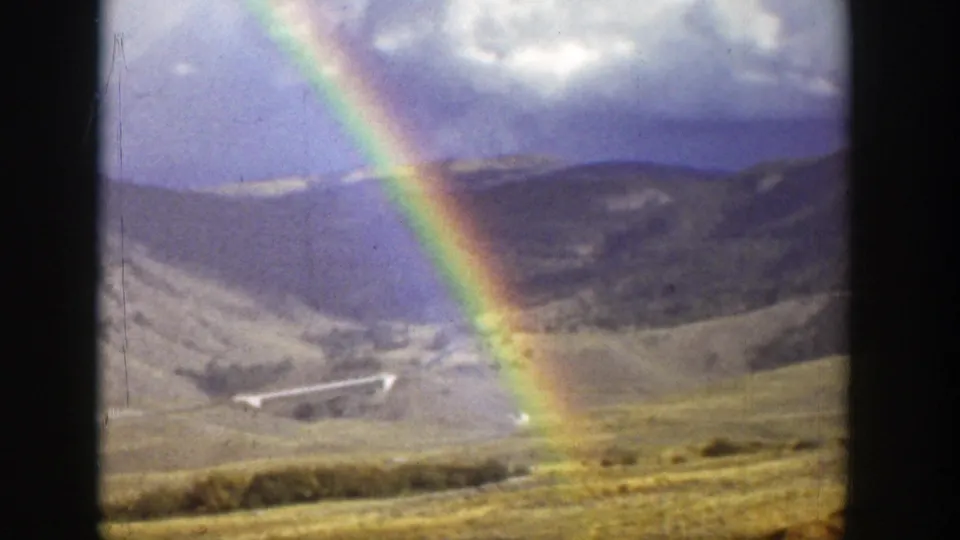 1939 Wyoming 8mm frame showing a rainbow arcing over dry hills, cloudy sky, scrubland, and a small distant bridge or road feature.