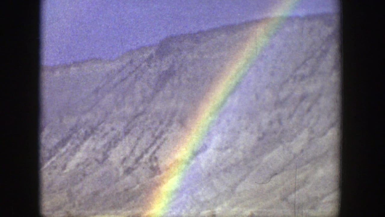 1939 Wyoming 8mm frame with a bright rainbow cutting across a pale, rocky hillside beneath a blue sky.