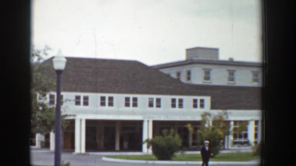 1939 Wyoming exterior of a large white building with dark roof, covered entrance, lamppost, shrubs, and a man crossing the foreground.