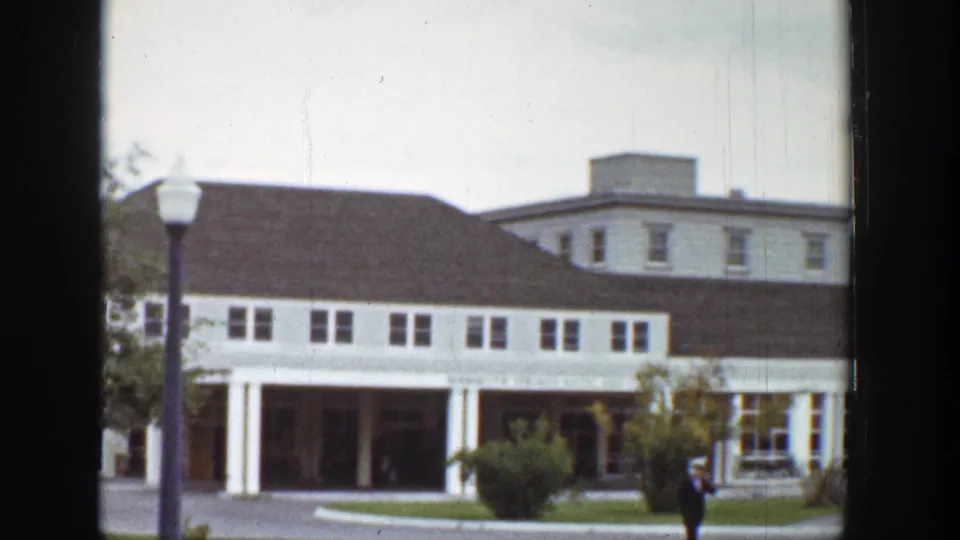 1939 Wyoming 8mm frame of a white multi-window building, dark roof, covered porch, lamppost, lawn, shrubs, and one man nearby.