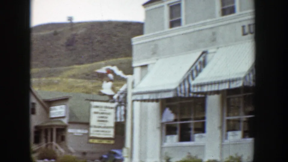 1939 Wyoming home-movie view of storefront windows, striped awnings, a roadside sign, parked car, and hillside background.