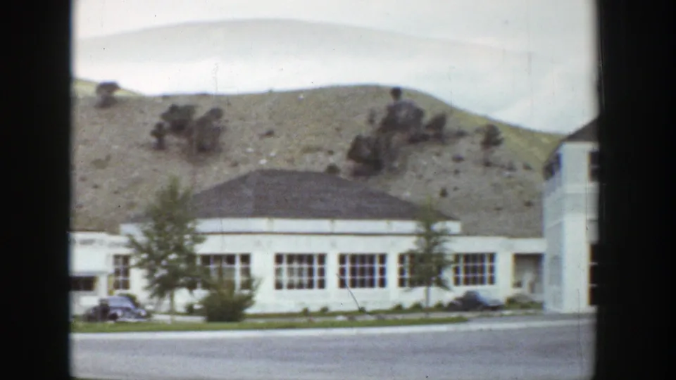 1939 Wyoming roadside view with a low white building, parked cars, small trees, paved road, and a barren hill behind it.