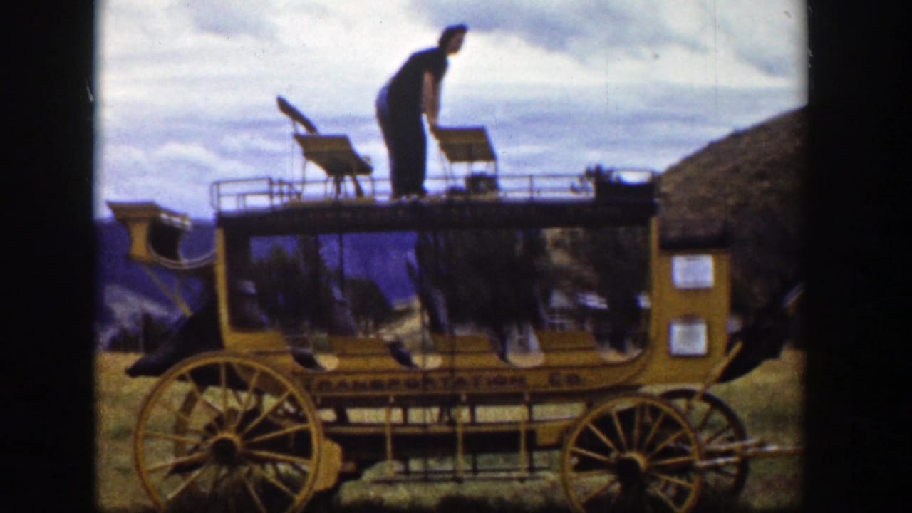 A man stands bent forward on the top of the yellow carriage, with roof seats, large wheels, hills, and cloudy sky visible.