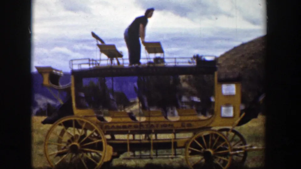 A man stands bent forward on the top of the yellow carriage, with roof seats, large wheels, hills, and cloudy sky visible.
