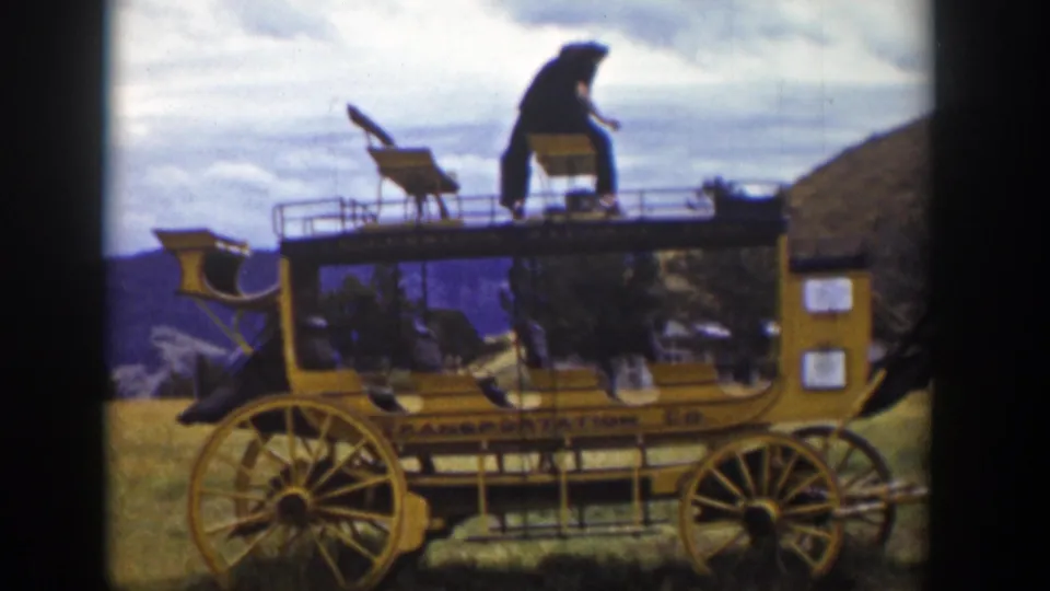 The man sits or crouches on the upper bench of the yellow stagecoach-style carriage, with hills and cloudy sky behind him.