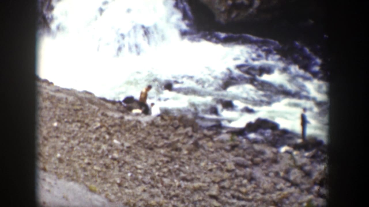 1939 Wyoming home-movie frame showing two distant people on a rocky bank near fast whitewater and a small waterfall.