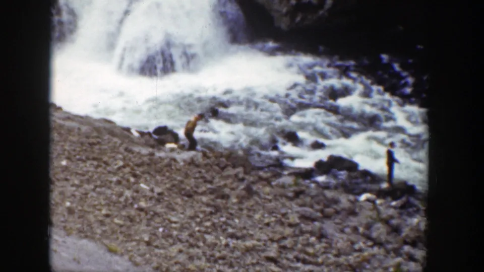 1939 Wyoming 8mm frame of a rocky shoreline, foaming river water, dark boulders, and two distant figures near the falls.