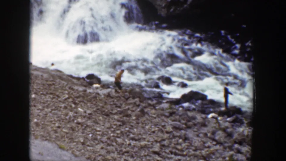 1939 Wyoming home-movie frame with two distant visitors on a rocky bank beside a bright, rushing waterfall.